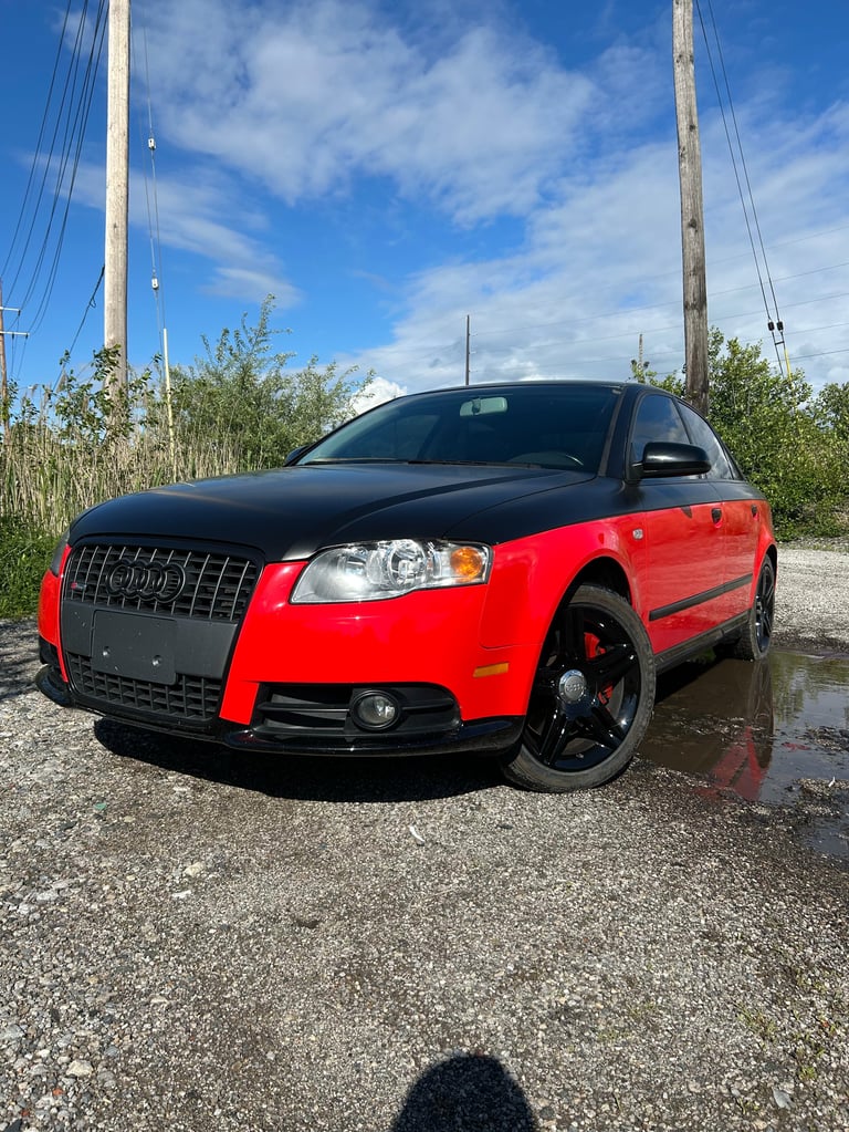 Red and black Audi sedan parked on gravel with tall poles and vegetation in background under blue sky