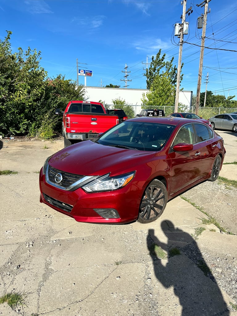 Red Nissan sedan parked on a lot with red pickup truck and other vehicles in the background
