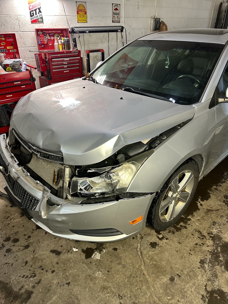 Damaged silver sedan in auto repair shop with hood and front end dismantled, exposing engine and headlights