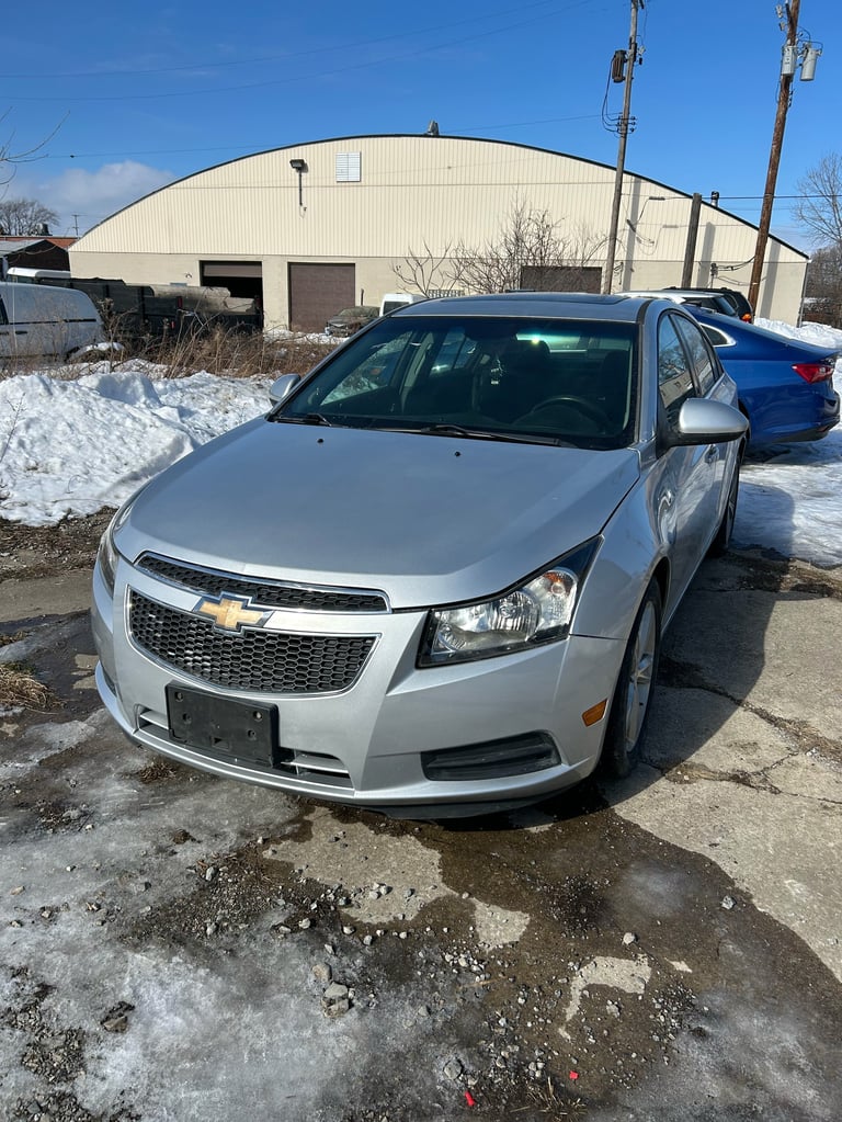 White Chevrolet Cruze parked in snowy lot with blue car and metal building in background