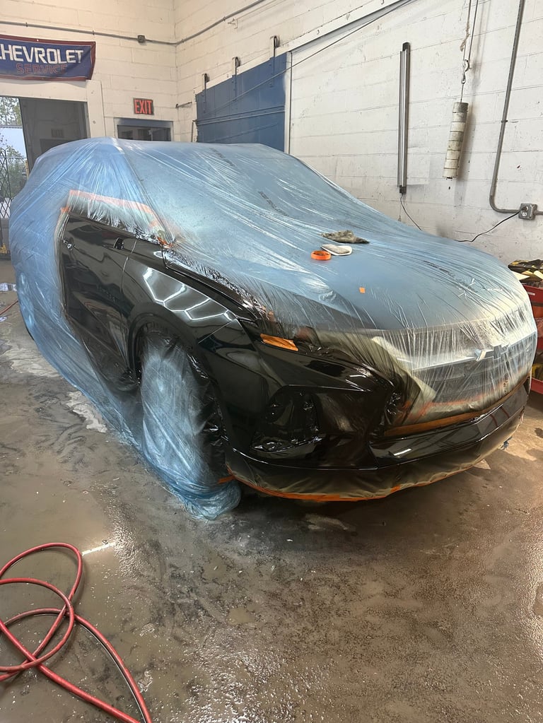 Black car wrapped in plastic sheeting in an auto repair shop garage with concrete floor and red hose nearby
