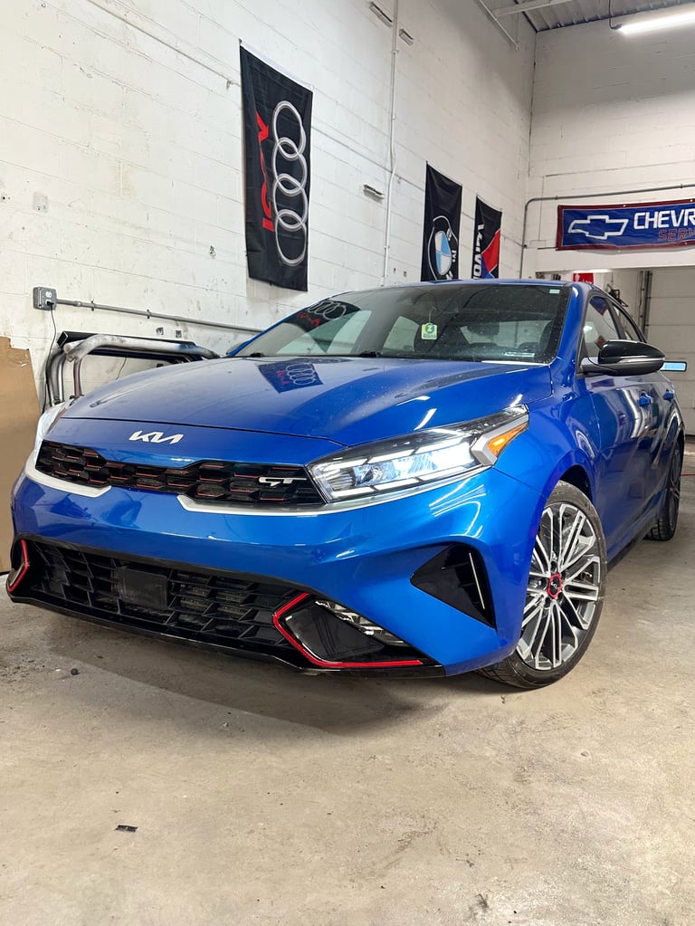 Blue Kia sedan on display in a garage with Audi and Chevrolet signage visible on the walls