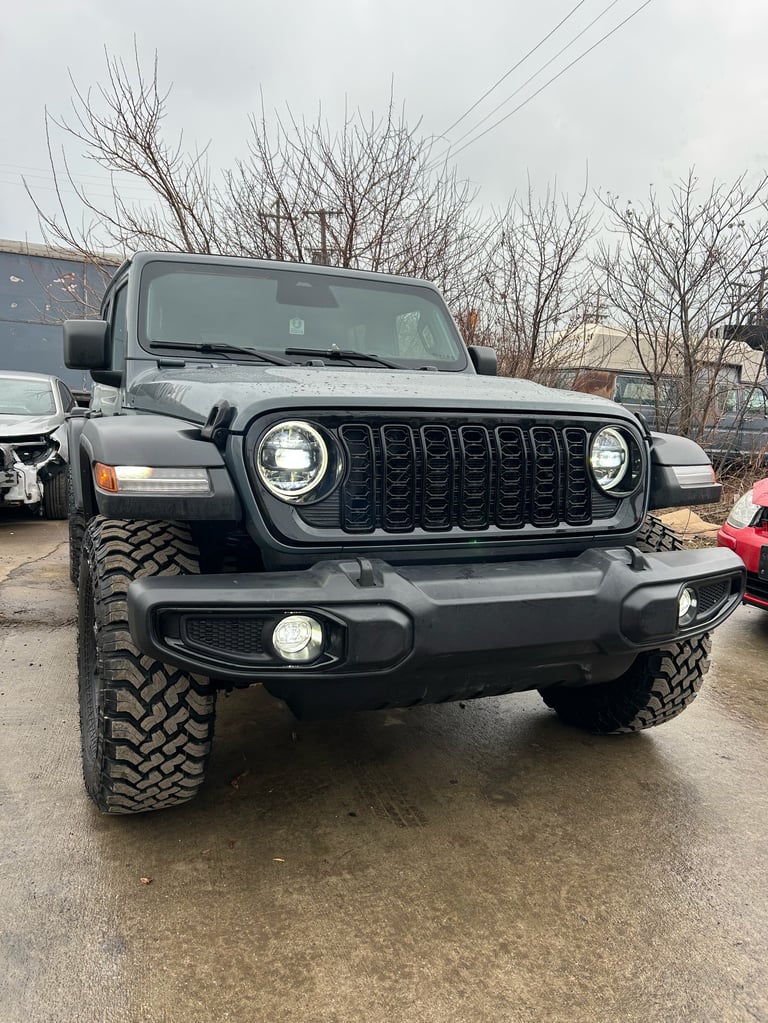Black Jeep Wrangler parked in lot with distinctive grille, headlights, and rugged tires on overcast day